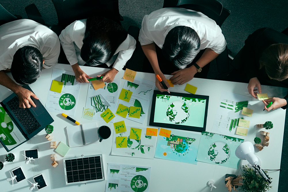 Top view of a team meeting around a table with clean energy reports, sustainability charts, a tablet, solar panel models and green planning notes.
