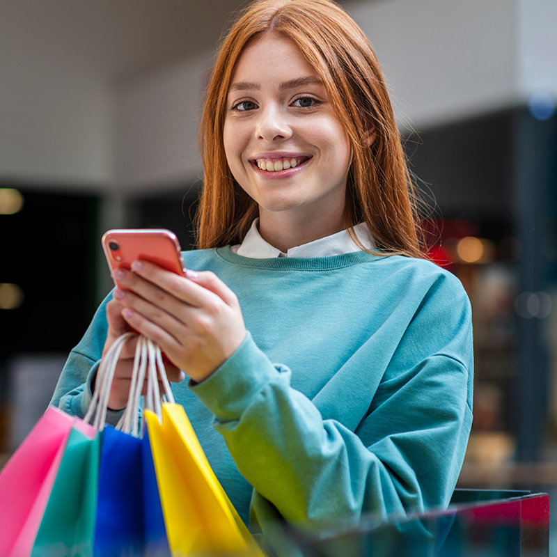 Smiling young woman holding shopping bags while using a smartphone in a shopping mall.