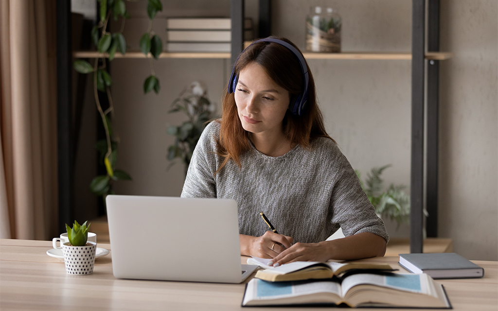 Woman focused on working at a laptop while taking notes in a notebook, seated at a desk with books and plants in a calm workspace.