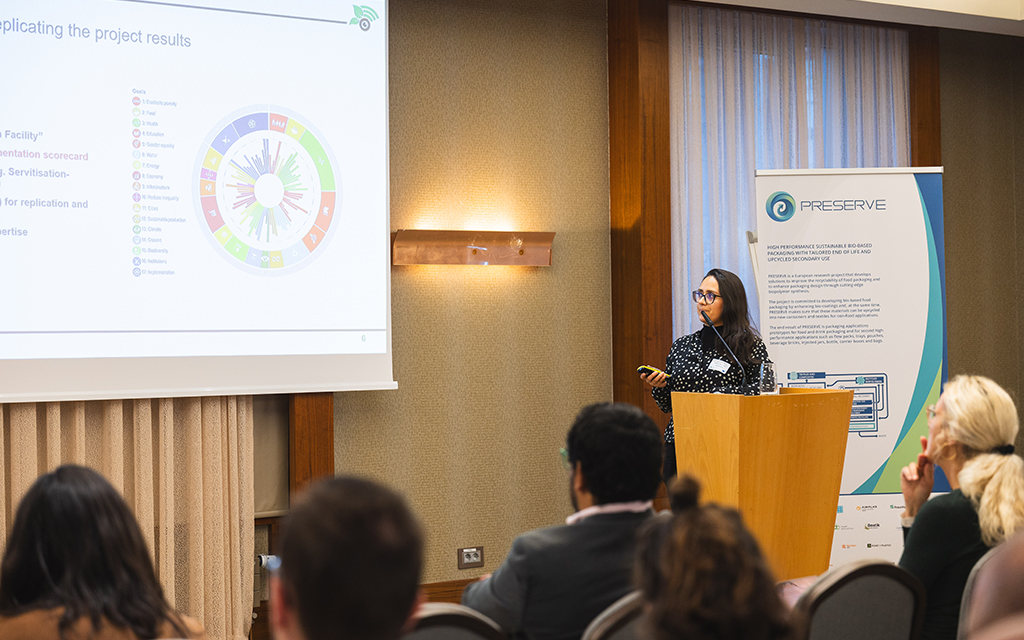 Conference speaker presenting project results at a podium, with a projected slide on screen and audience seated in a hotel meeting room.