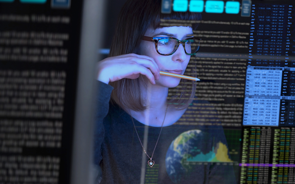 Woman wearing glasses reviewing computer code on a screen while holding a pencil.