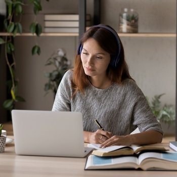 Woman focused on working at a laptop while taking notes in a notebook, seated at a desk with books and plants in a calm workspace.