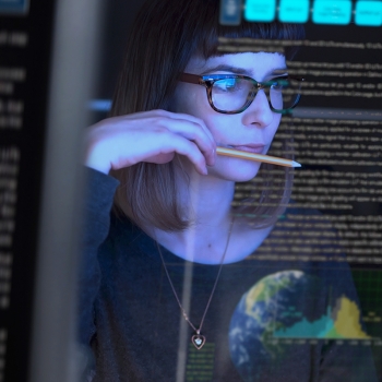 Woman wearing glasses reviewing computer code on a screen while holding a pencil.
